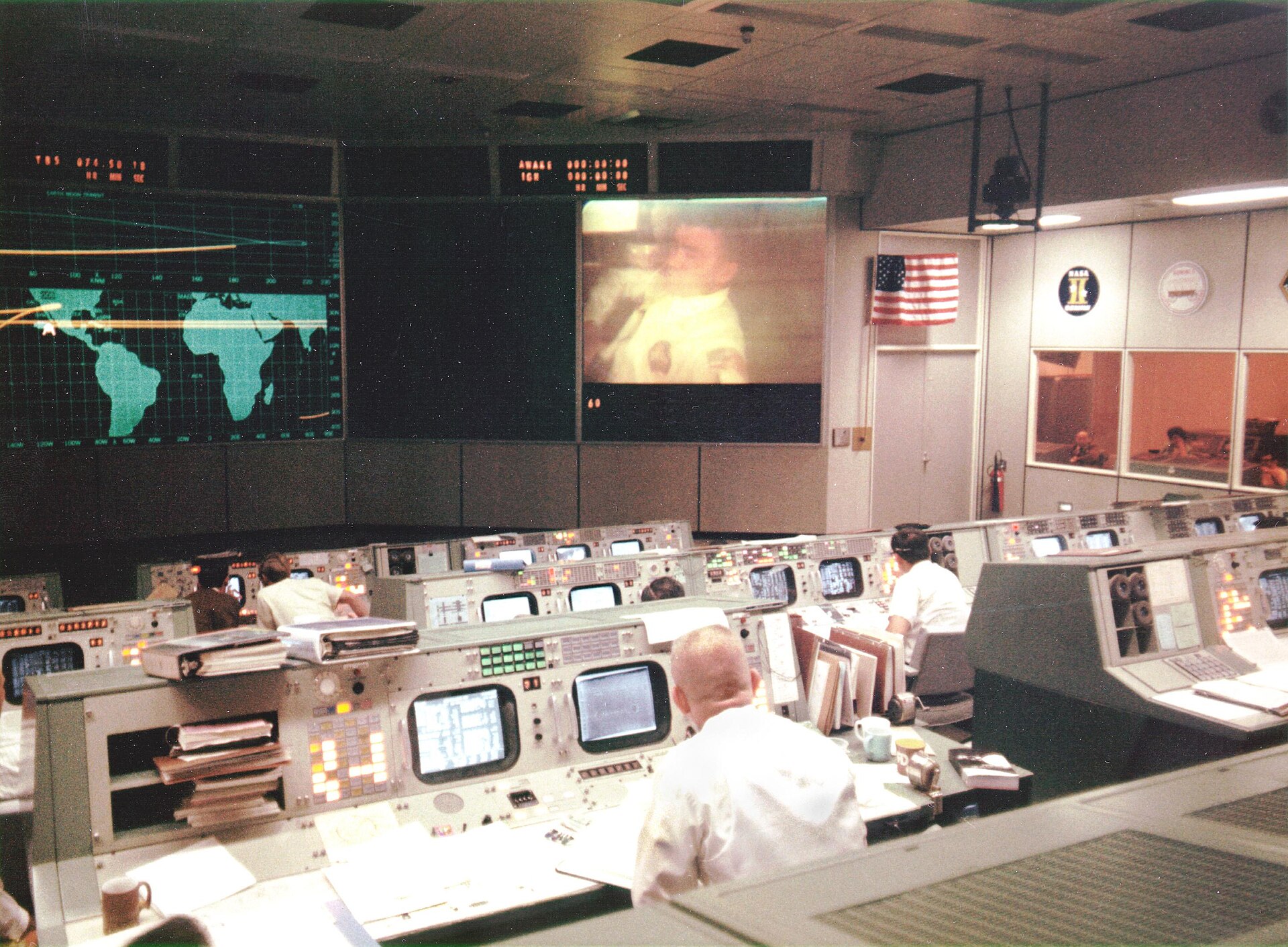 Flight controllers at their consoles in Mission Control during Apollo 13, with a world map and live TV feed on the wall screens
